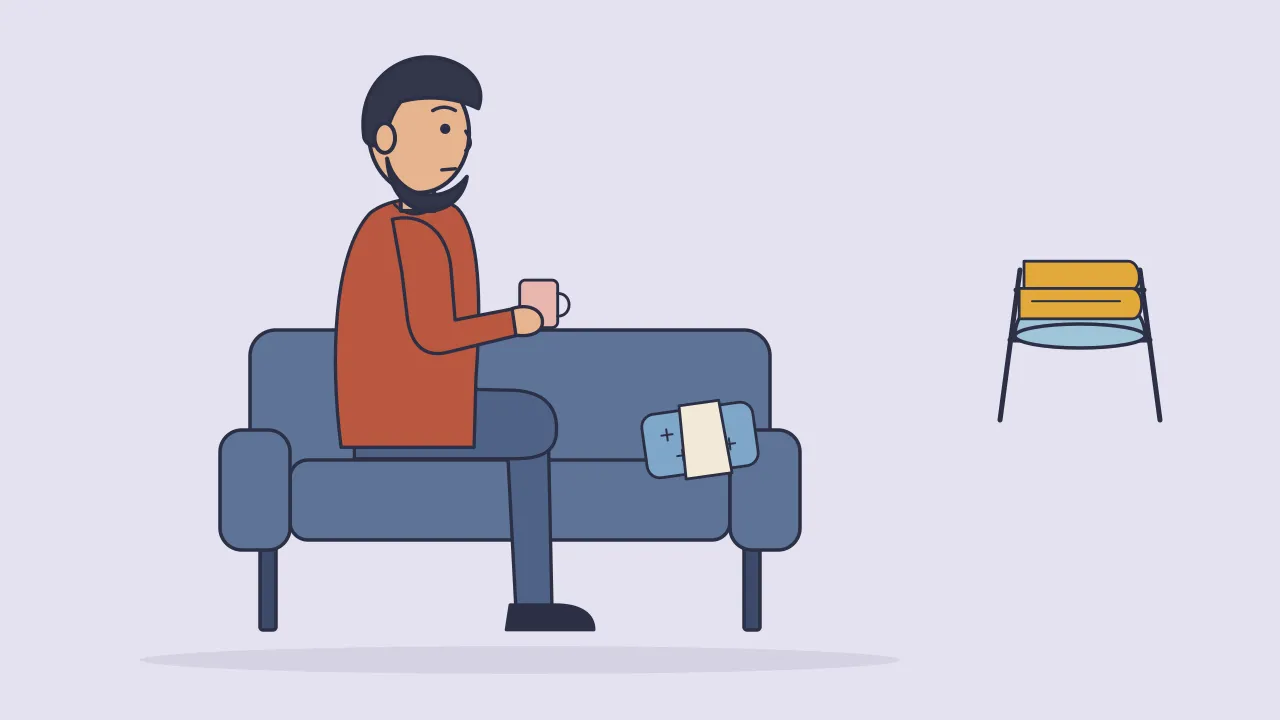 A person on a sofa holding a mug, with a wrapped cold pack on the cushion beside them and a sitz bath basin and towel drying on a rack
