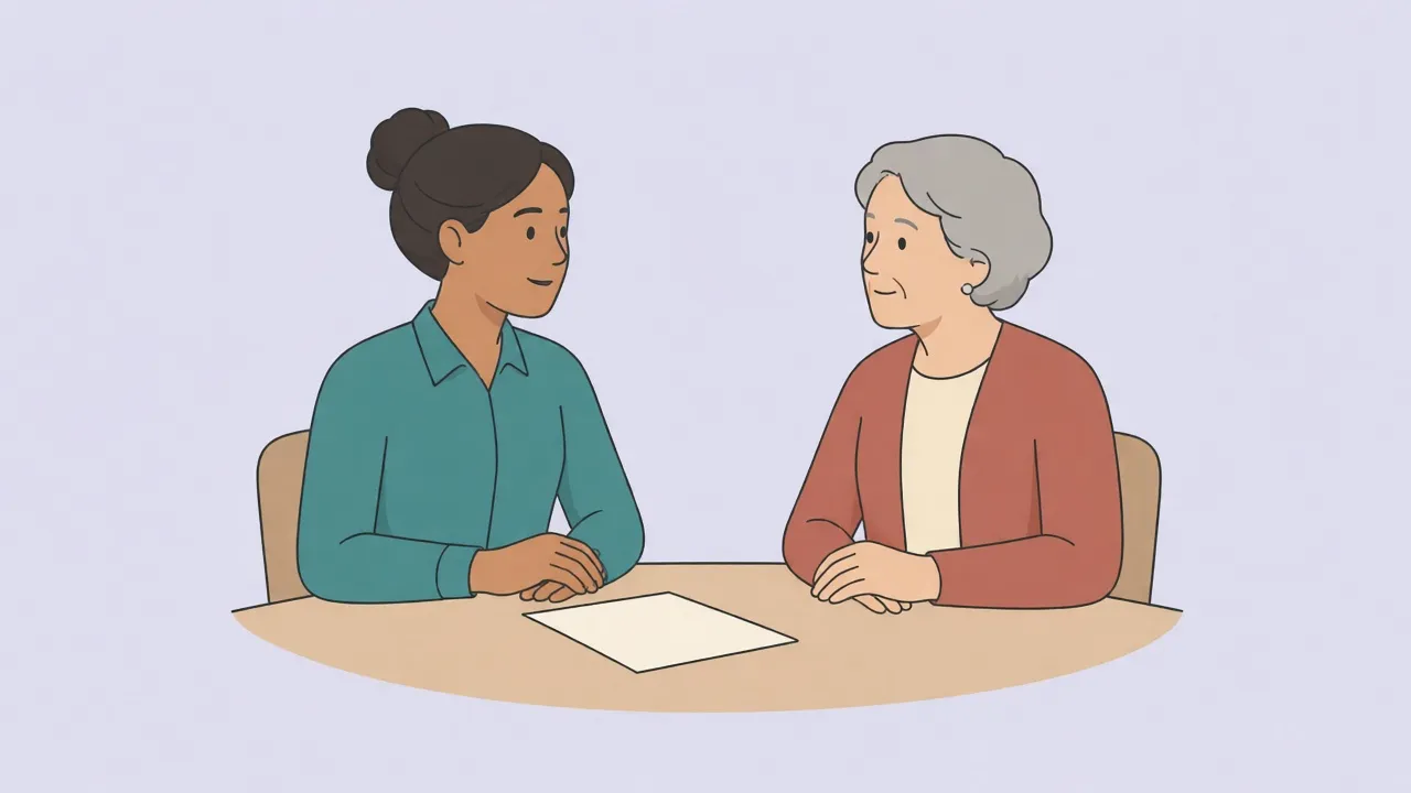 A clinician in a teal shirt sits across a small wooden table from an older patient in a rust cardigan, both fully clothed and talking, with a blank sheet of paper on the table